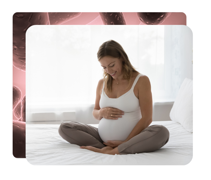 En gravid kvinde sidder med korslagte ben på en seng og holder om sin mave og smiler, billedet ligger ovenpå en lyserød baggrundsgrafik med bakterieformer.